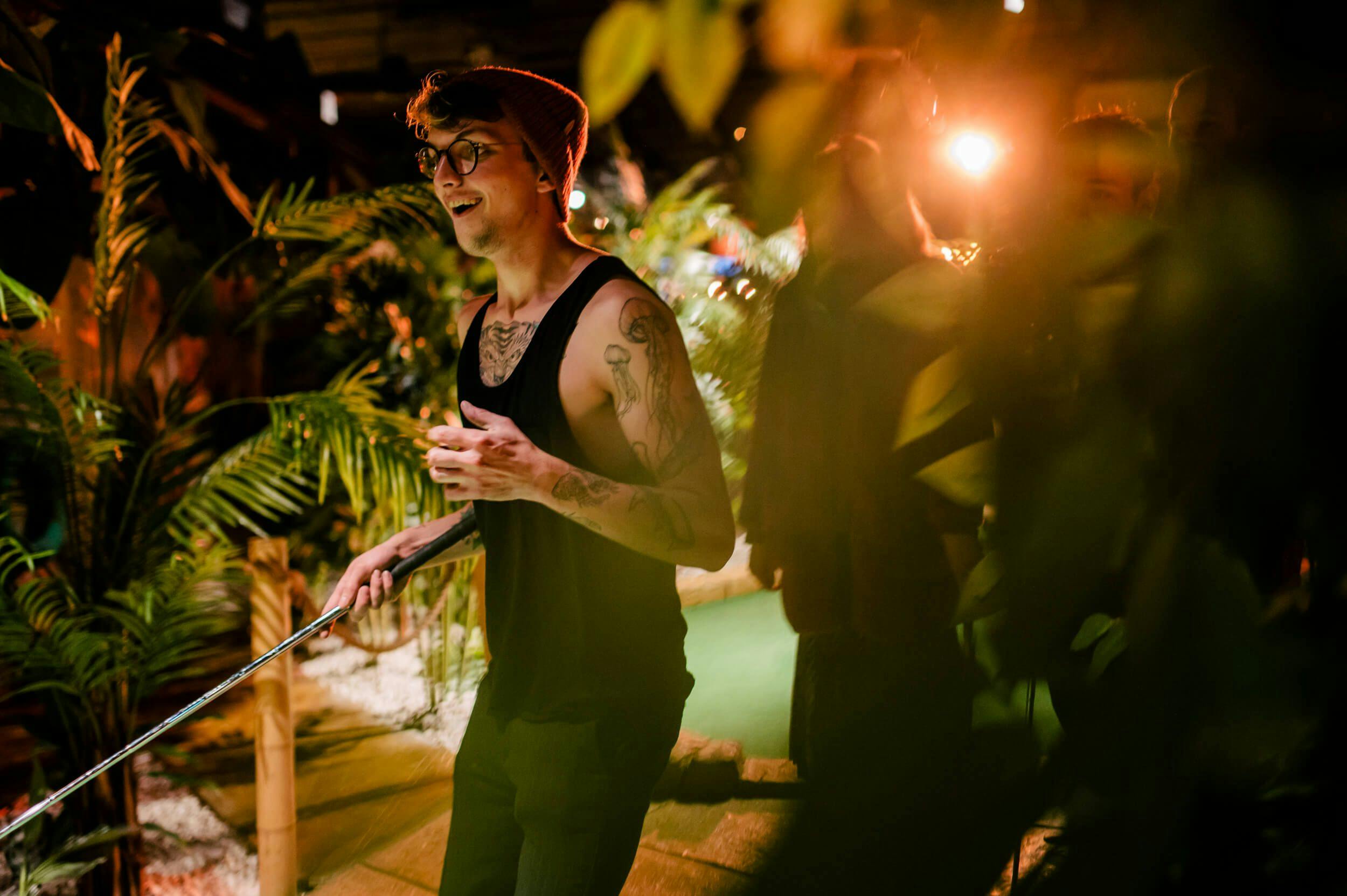 Image of a man in a black vest wearing a beanie, glasses and covered in arm tattoos walking through the Treetop jungle holding a mini golf club while his friends trail behind.  