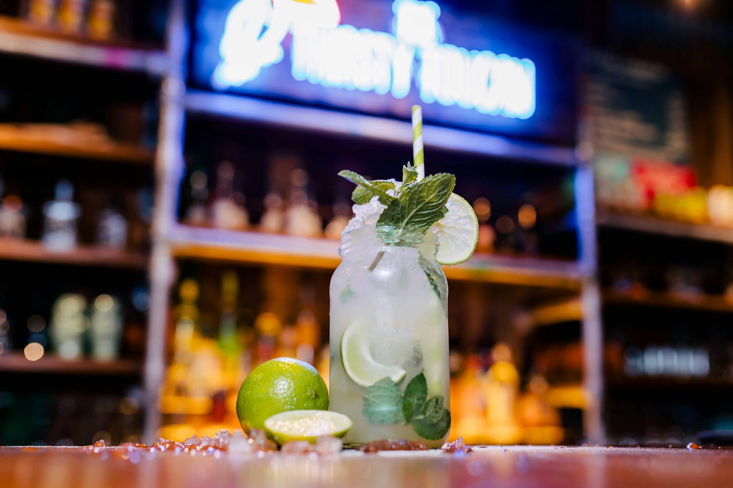 Image of a fruity lime and mint cocktail on the bar of the Thirsty Toucan; a fluorescent light beams blue in the background.