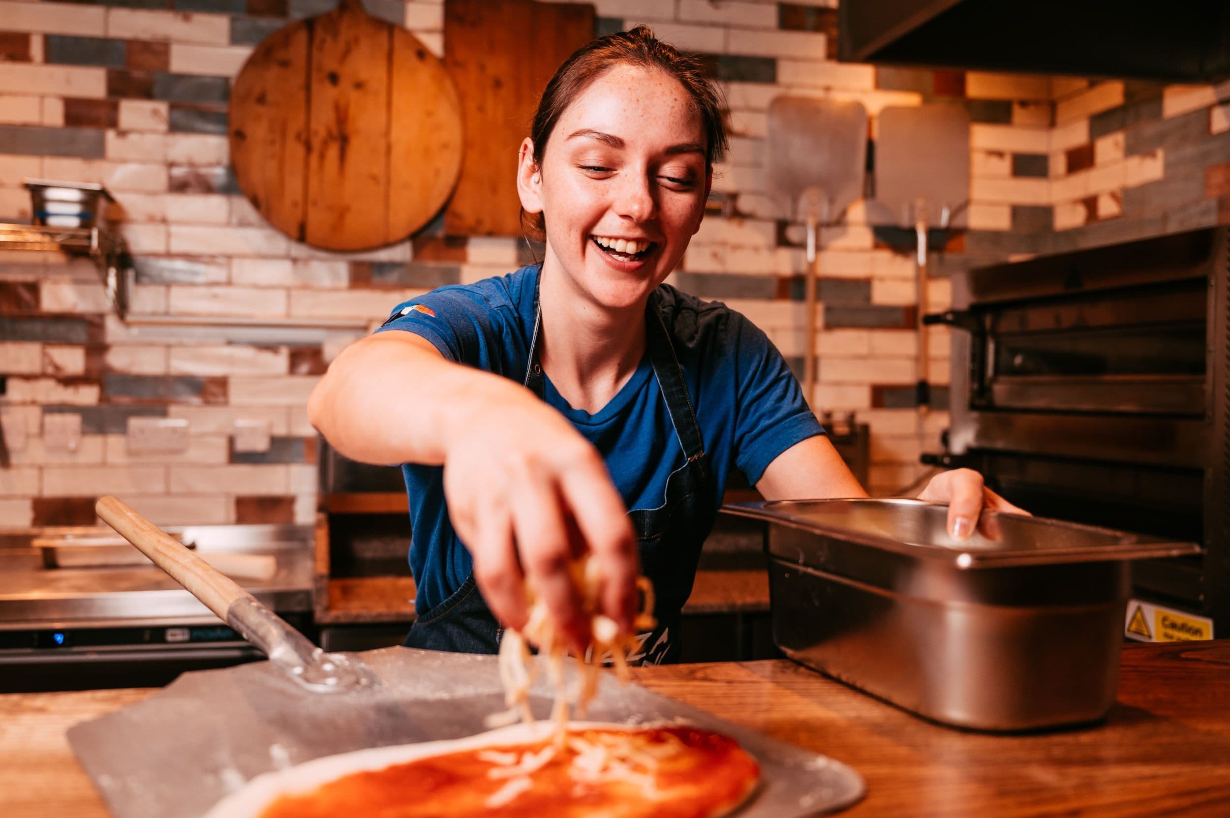 Girl with her hair tied back and a blue t-shirt sprinkling cheese over a raw pizza base while smiling.