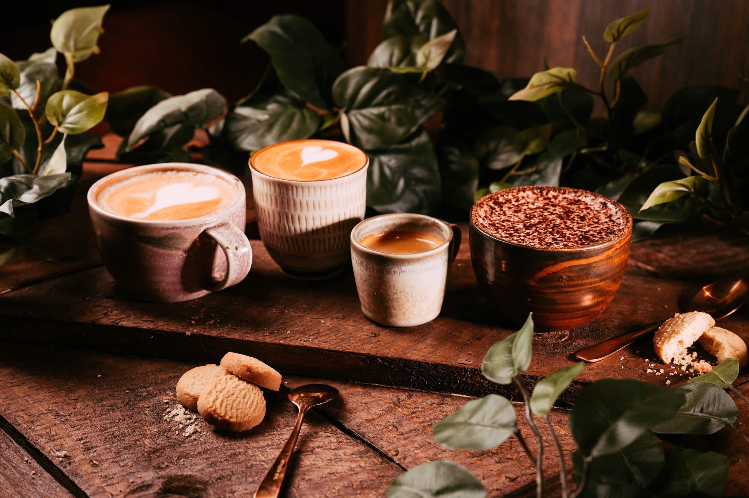 Tray of delicious coffees, including a cappuccino, latte, espresso shot and hot chocolate on a wooden board with biscuits and a spoon next to it.