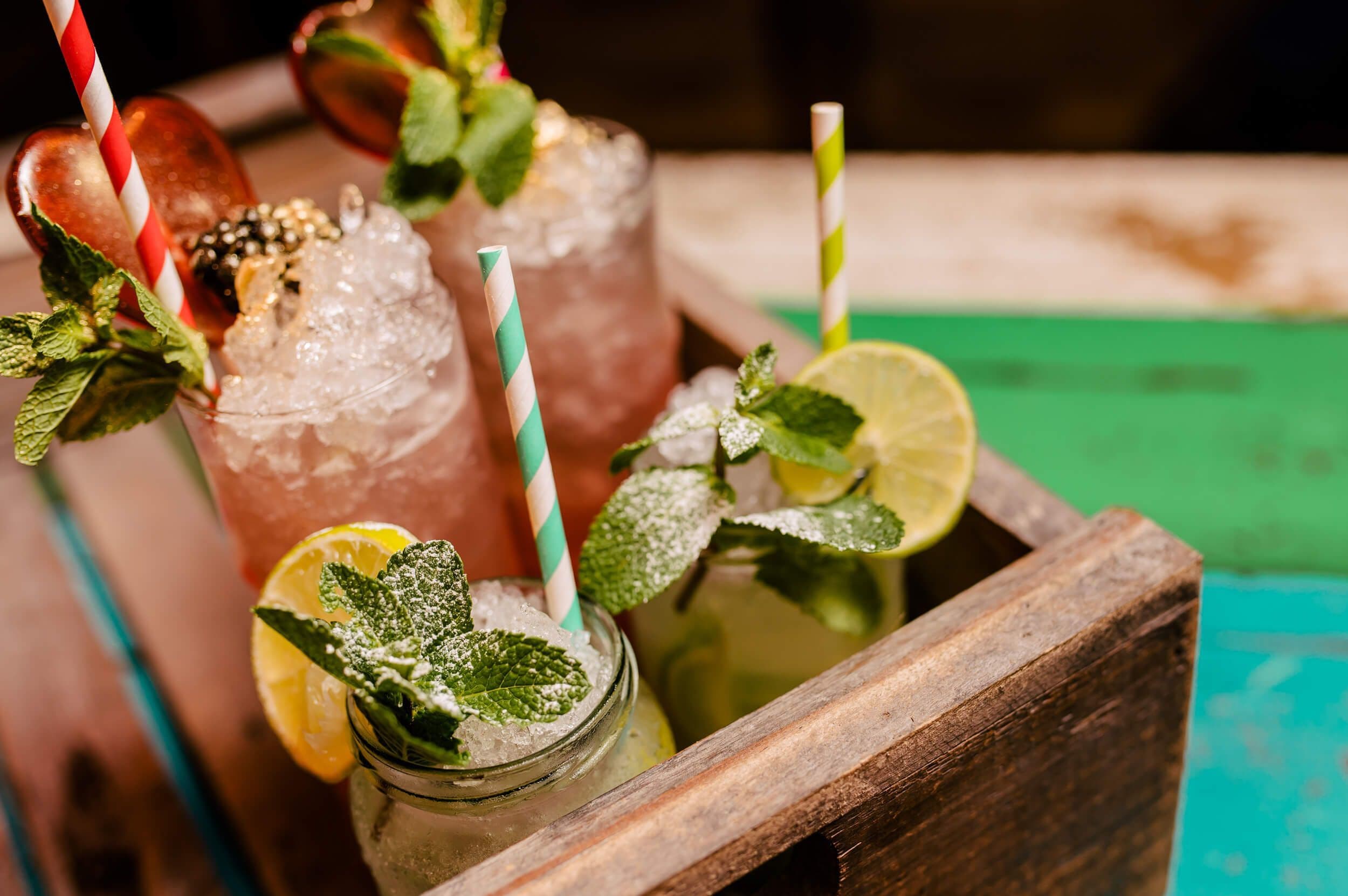 Image of tropical pink and green cocktails, garnished with mint, lemon and frosted by powdered sugar in a wooden crate.