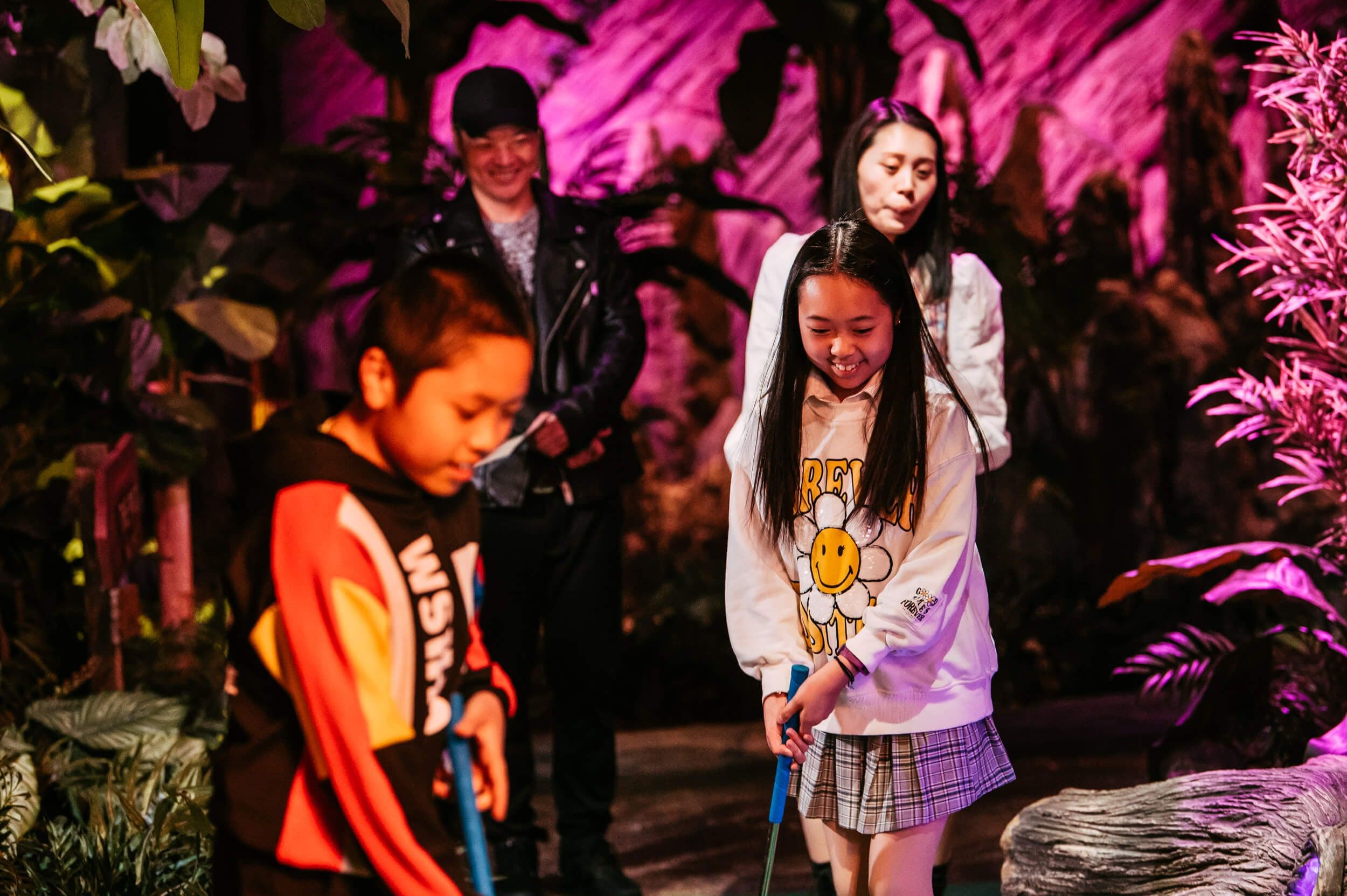 Young family play mini golf together on the Tropical Trail with bright pink light cast over the course. 