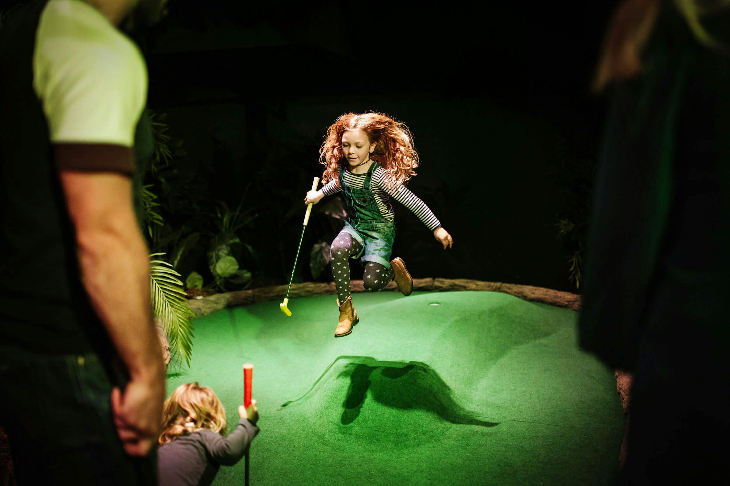 Young girl jumping over a mini golf green hurdle, with a club in one hand and her hair flying. 