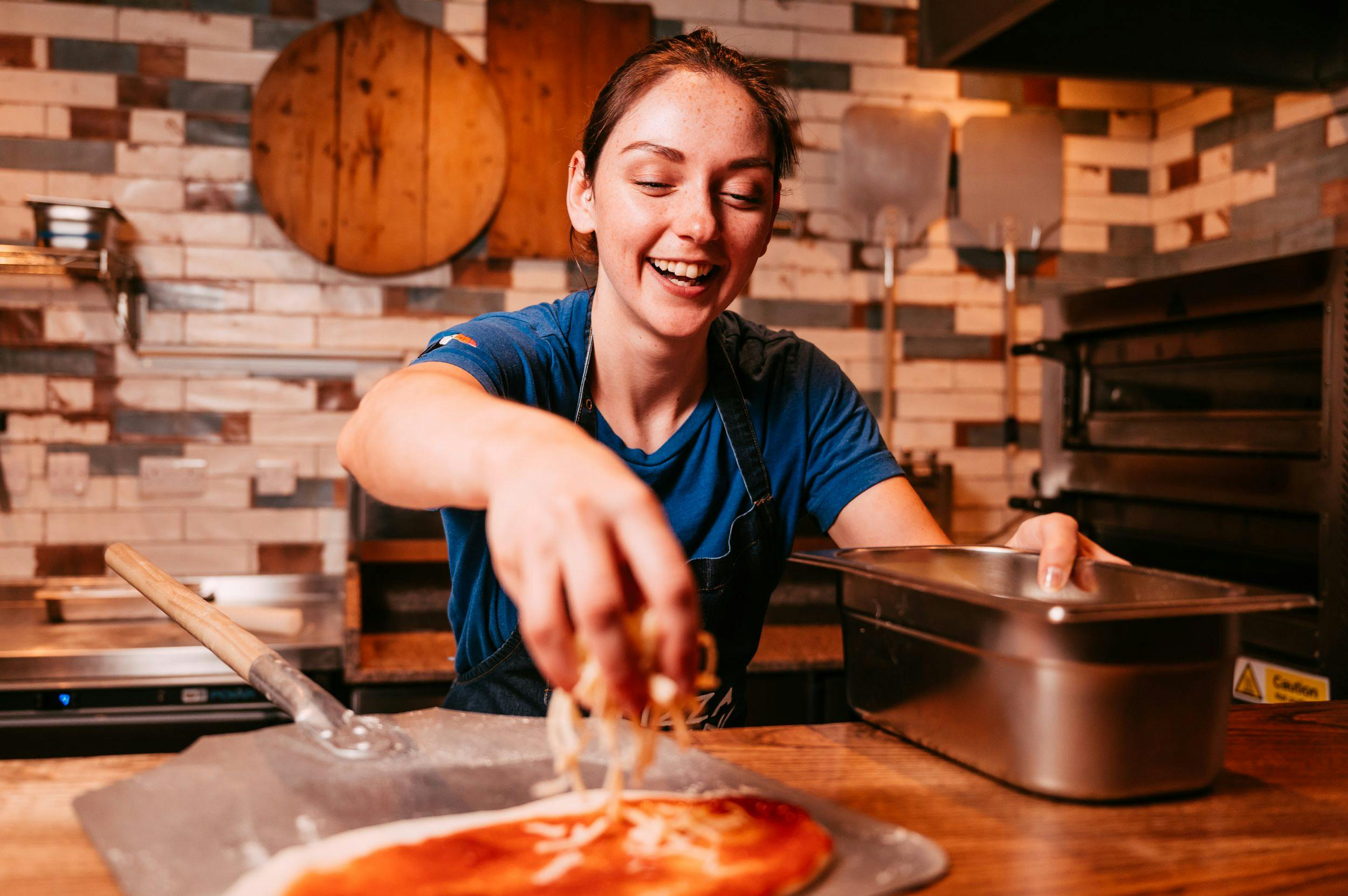 Girl with her hair tied back and a blue t-shirt sprinkling cheese over a raw pizza base while smiling. 