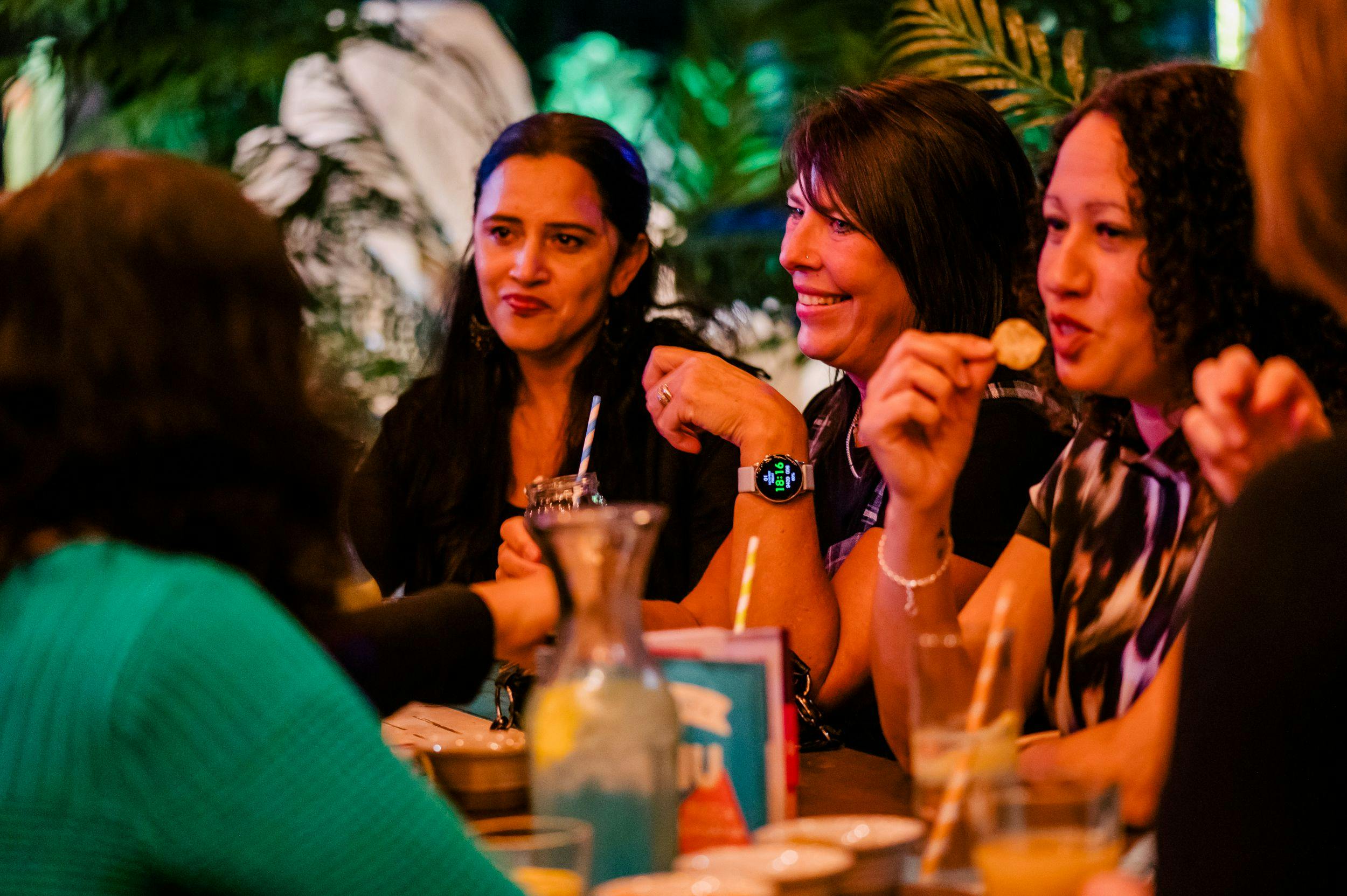 A group of women sit at a table in the market surrounded by cocktails and food, eating as they talk and laugh. 