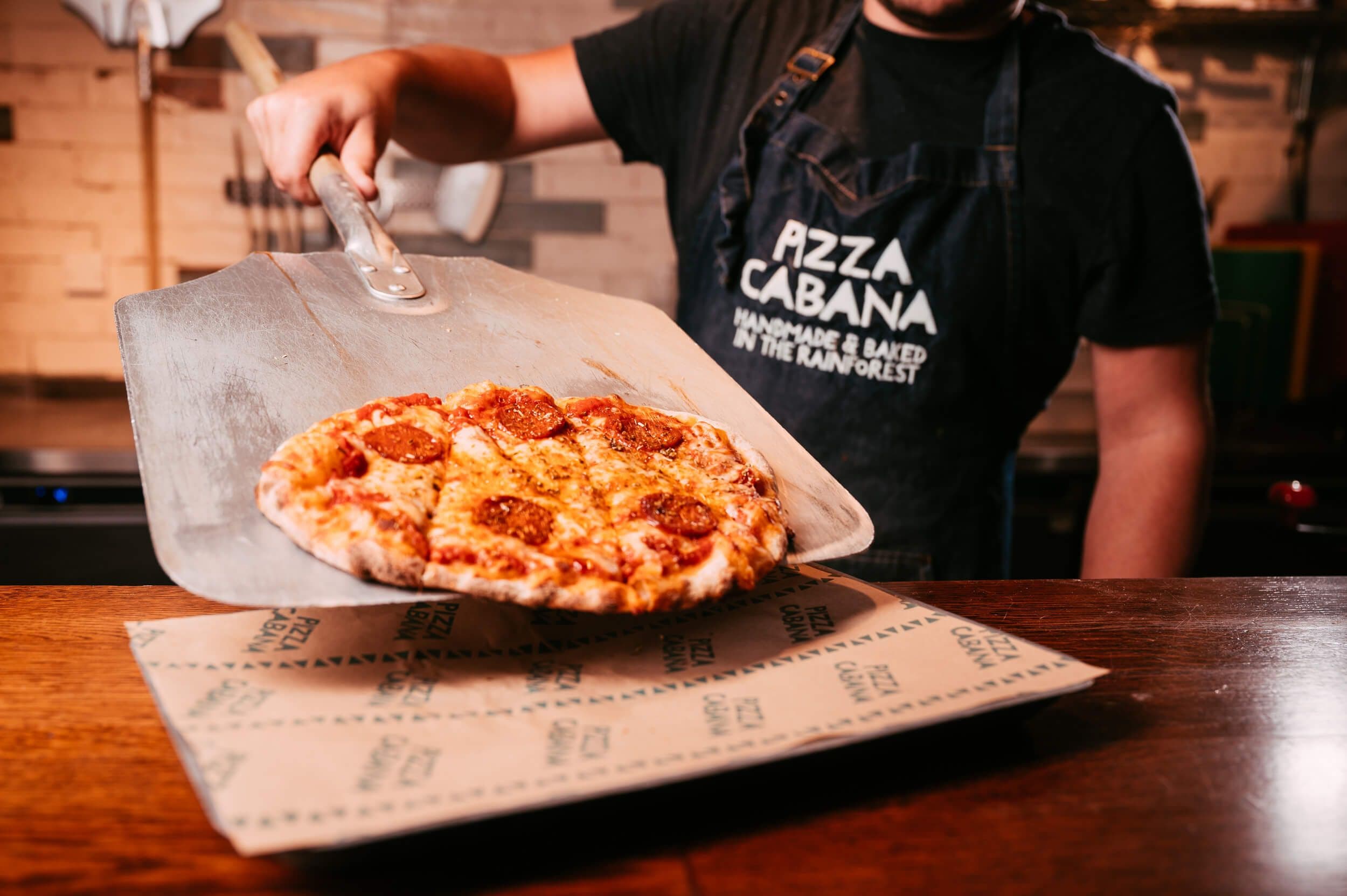 Man wearing a Pizza Cabana apron sliding a freshly wood fired pizza with pepperoni onto a tray.  