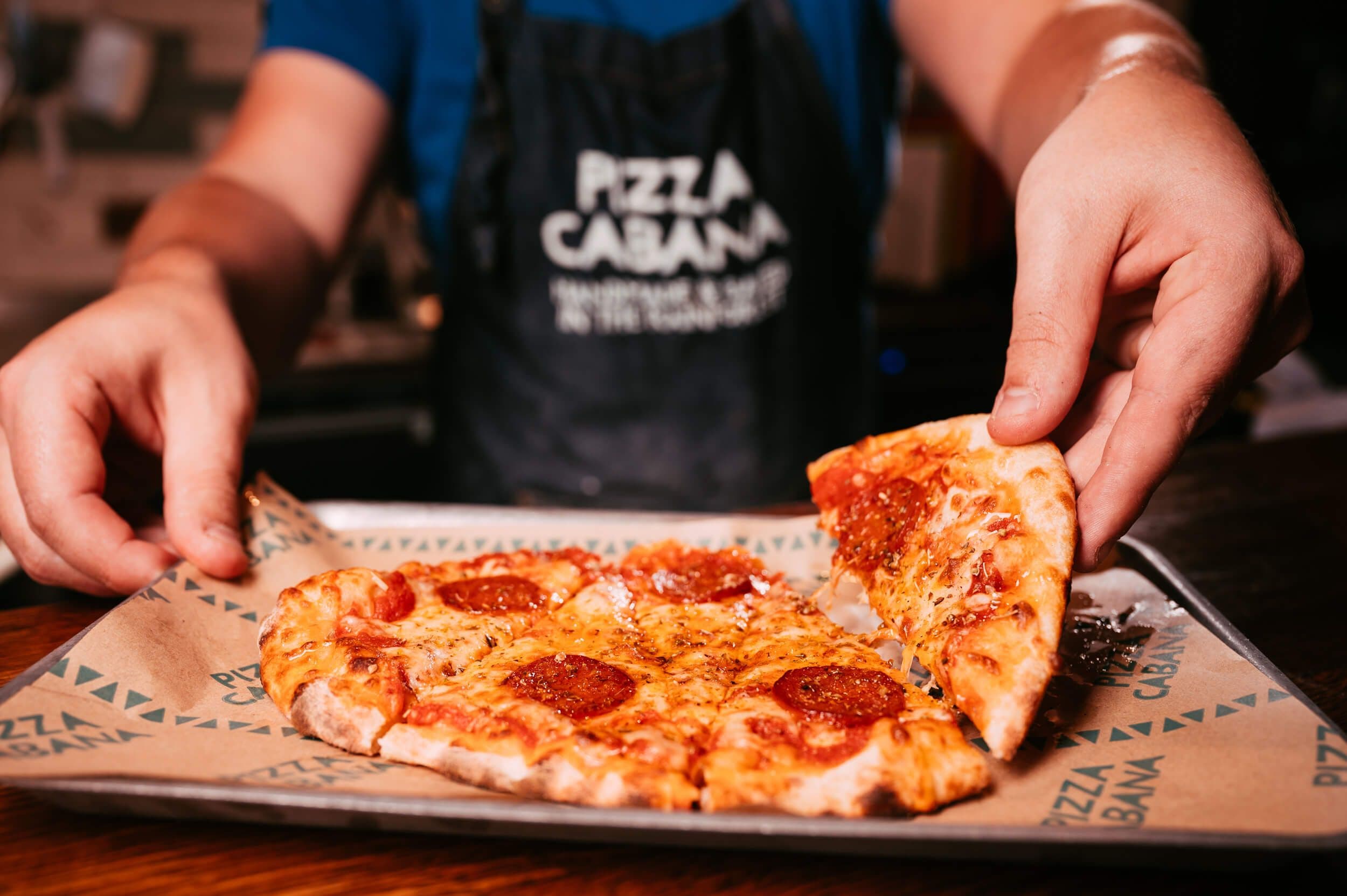 Image of a man lifting one of the Pizza Cabana slices on a classic Treetop tray, while wearing a Pizza Cabana staff apron. 