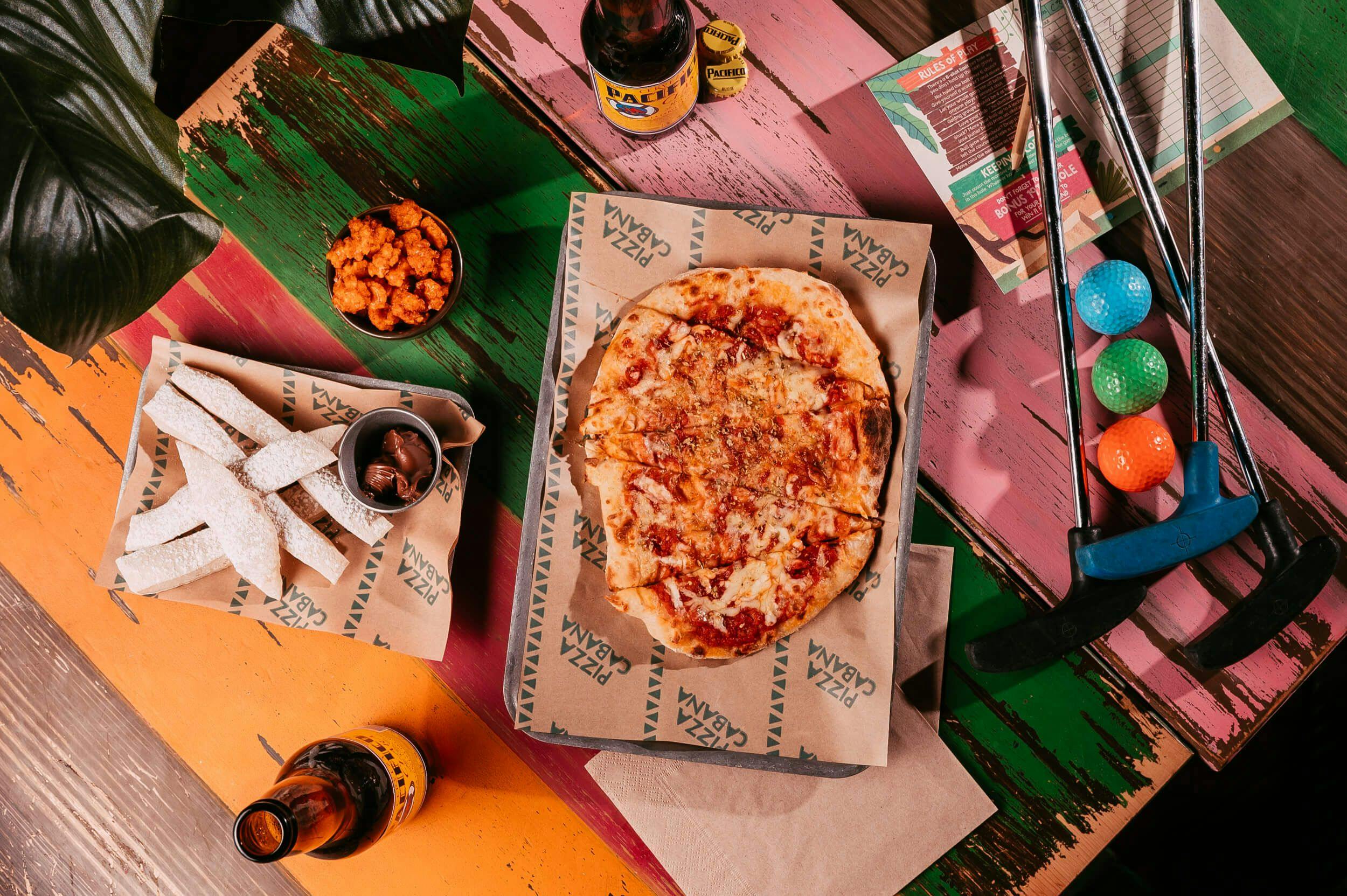 Yellow, red, green and pink striped wooden table covered in treats from the Pizza Cabana, like beer and wonky, handmade pizza and dough sticks. 
