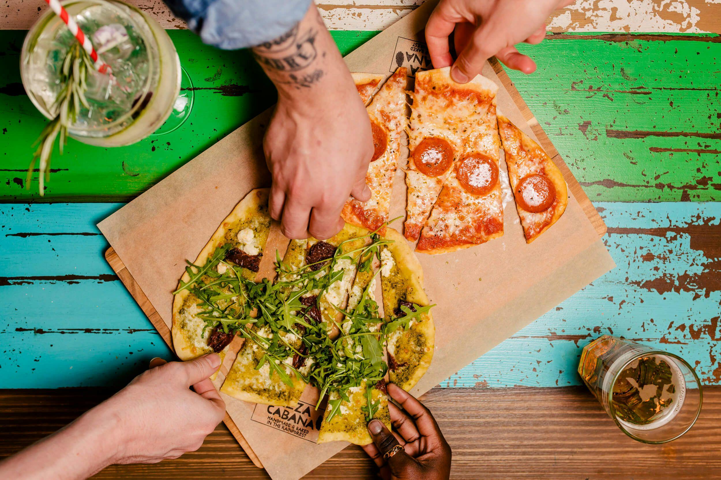 A birds-eye-view of a striped blue, brown and green table with two Pizza Cabana wonky, oval pizzas in the centre being enjoyed by two people.