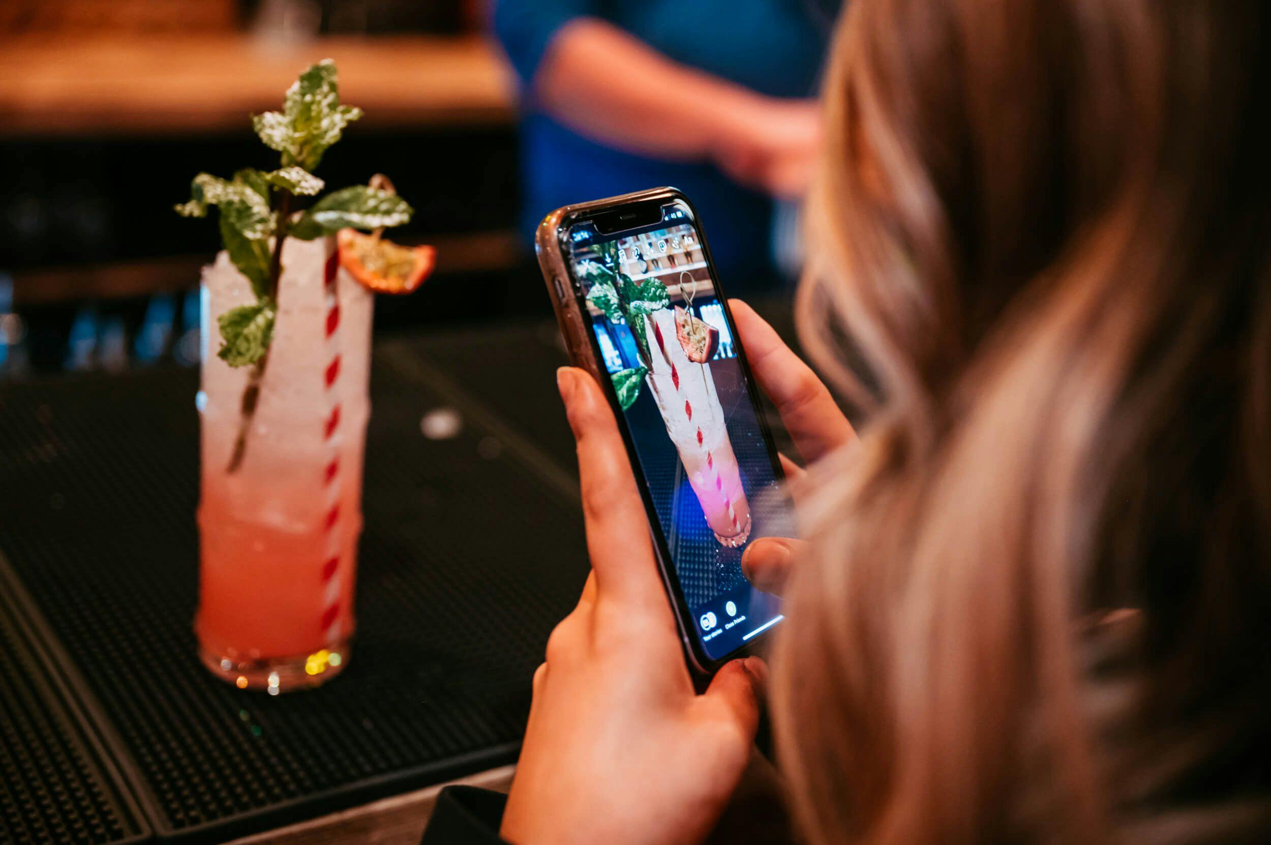 Image of a women with blonde hair taking a photo of a bright pink cocktail on her phone, zoomed in on the garnish. 