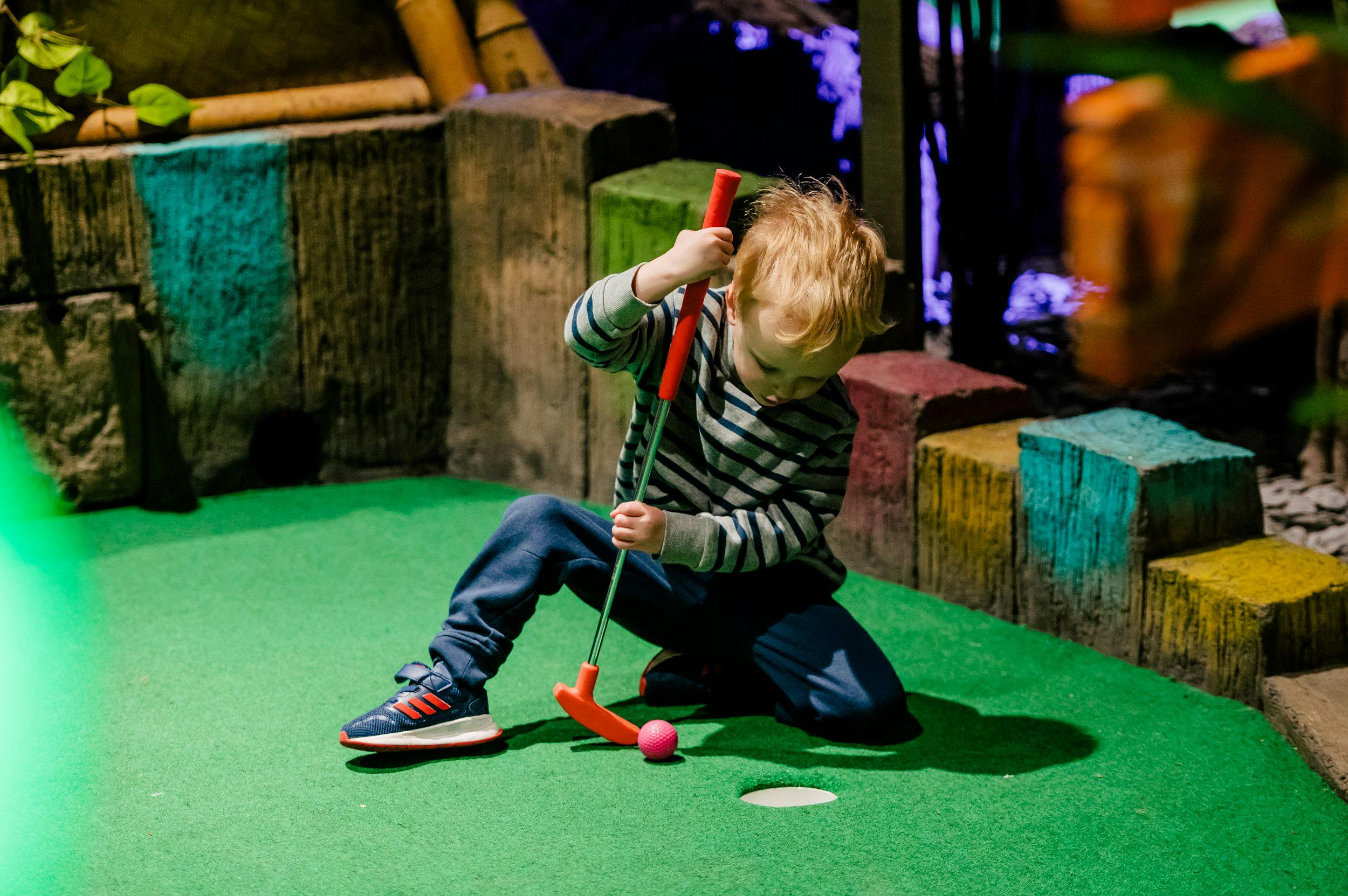 A small toddler kneeling down to hit the golf ball.
