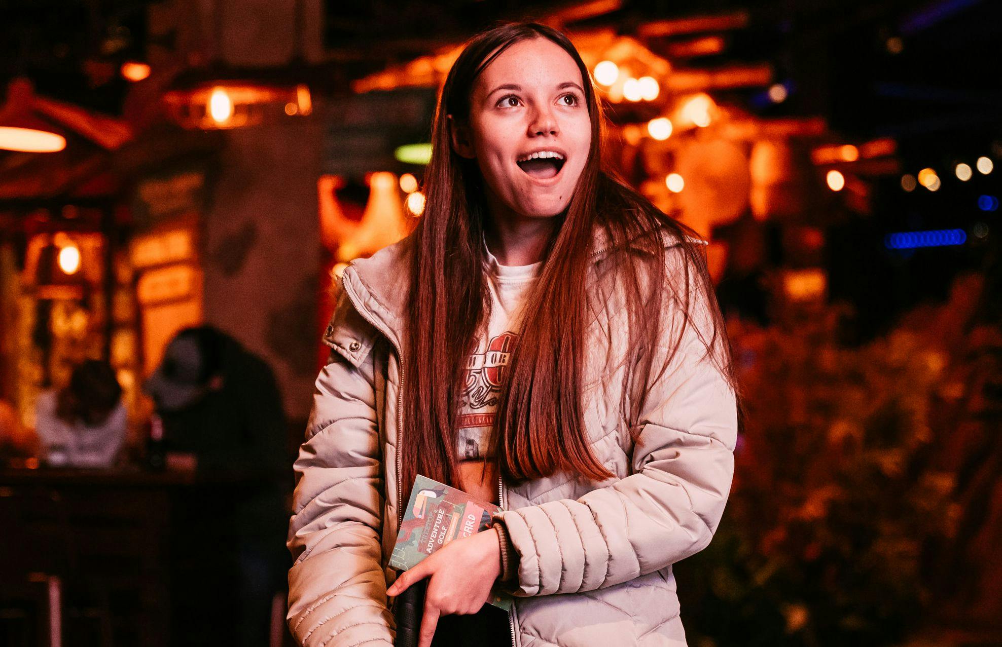 Happy girl with long hair looks around with her mouth open in awe at the Treetop adventure golf sights, wearing a nude puffer jacket. 